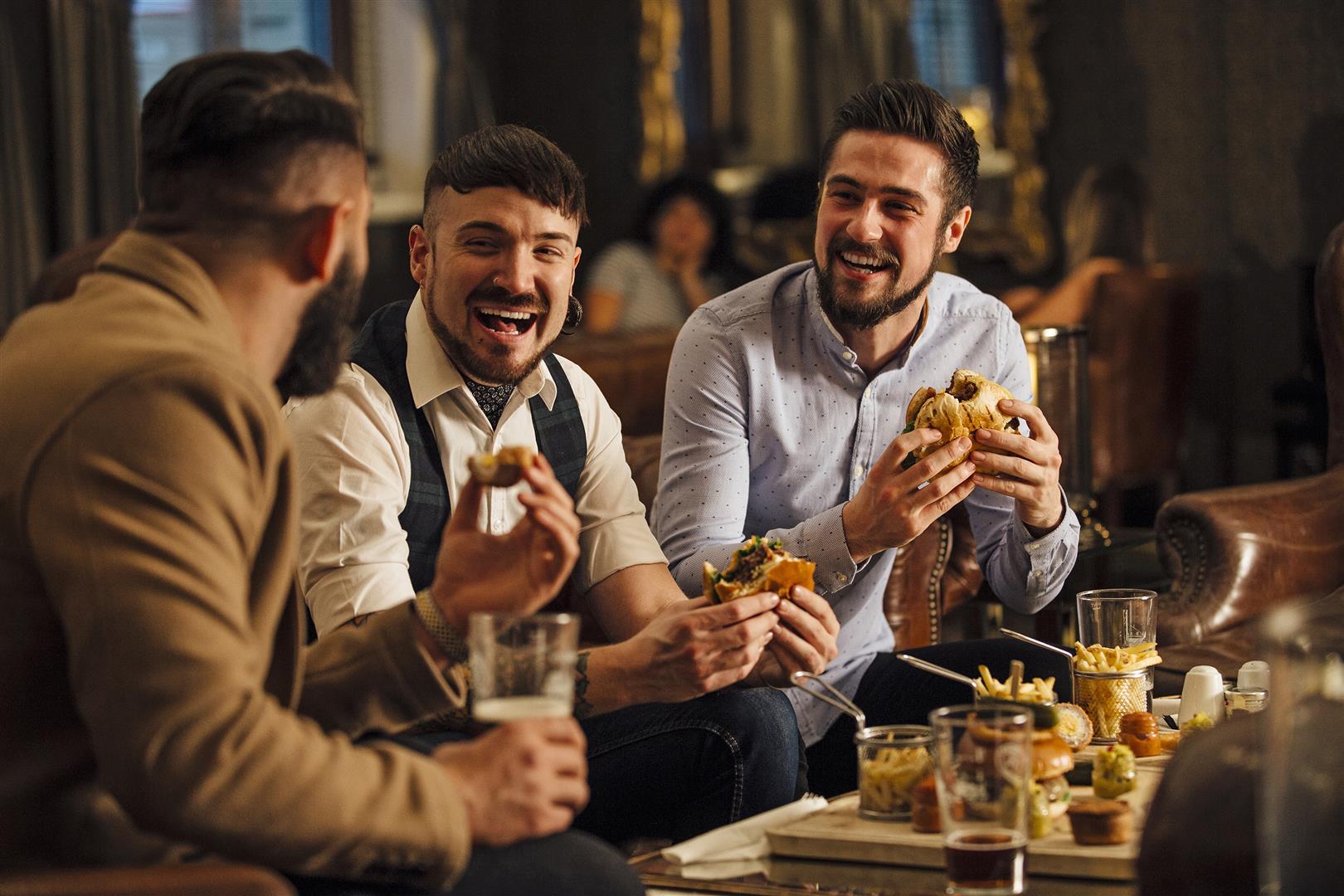 Three men are sitting together in a bar/restaurant lounge. They are laughing and talkig while enjoying burgers and beer.